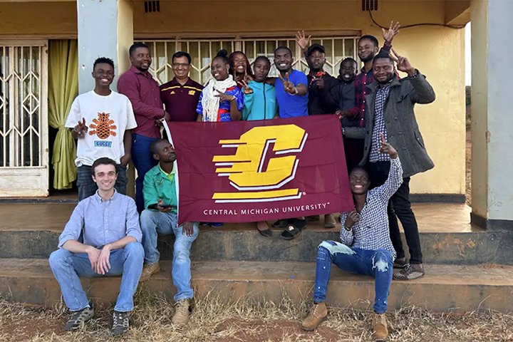Professor Prakash Adhikari (back row, third from left, wearing maroon CMU shirt and glasses) and CMU student Brady Whalen (front row, left wearing blue shirt and jeans) with global classroom students in Dzaleka holding a CMU flag.