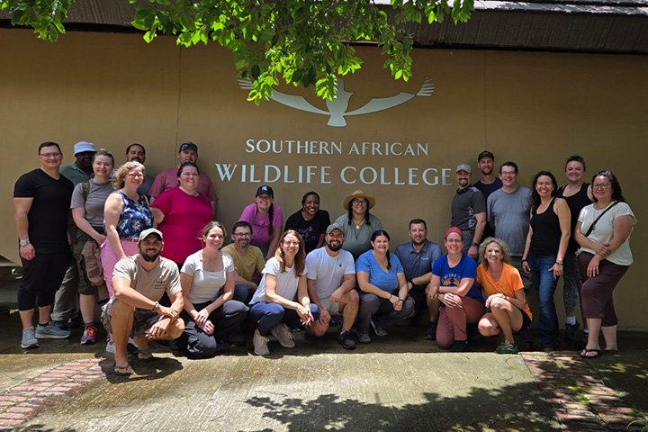 Andrea LaFontaine poses with a group of people kneeling and standing below a sign for the Southern African Wildlife College