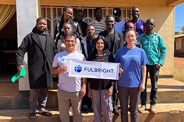 Prakash Adhikari, Niraja Adhikari, and fellow Fulbright Scholar Molly Dunn stand on steps holding a Fulbright sign with Global Classroom students on steps behind them.