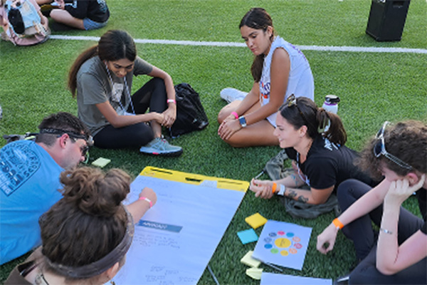 NextGen students sitting together on a green turf field writing ideas on a large pad of paper laying on the ground between them.