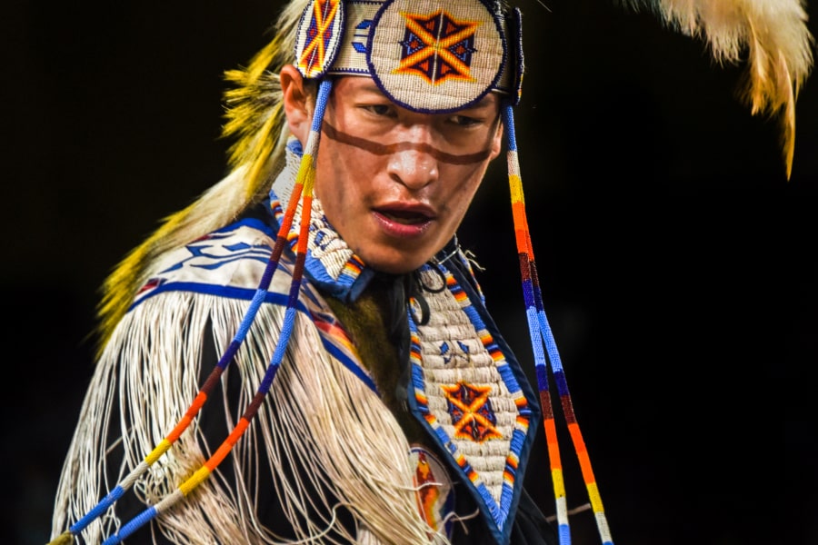 A male Native American dancer in traditional regalia performs at a Pow wow. He wears a beaded headband and chest piece with geometric patterns in orange, blue and white, along with long fringe and braided ribbons.