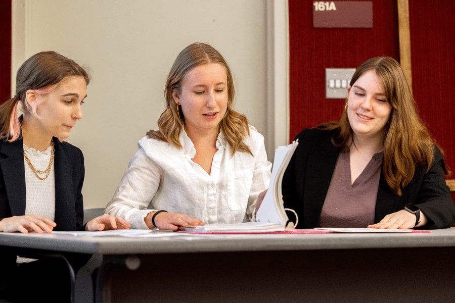 Three female pre-law students sitting at a table looking at a binder of papers.