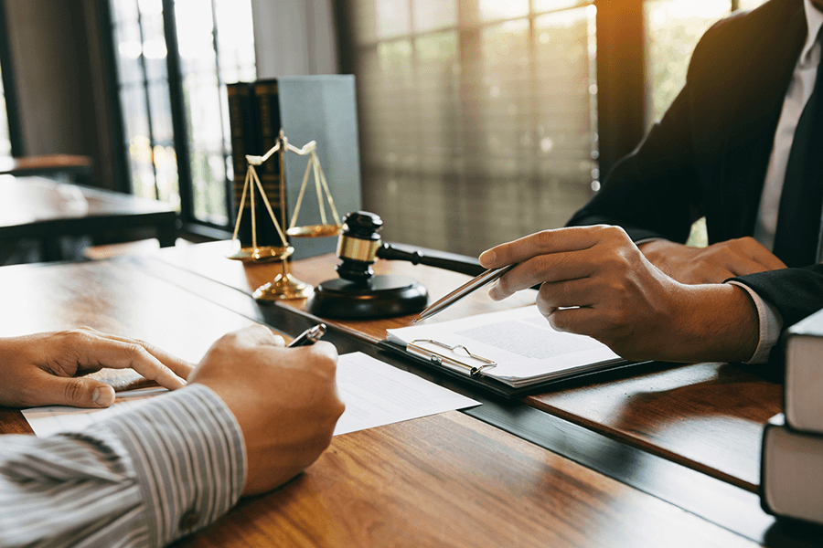 Two individuals sitting across from one another, a close up of their hands with paper and pens in front of them. In the background on the table next to them sits a gavel, as well as a judicial gold scale, with windows and the sun shining into the room.