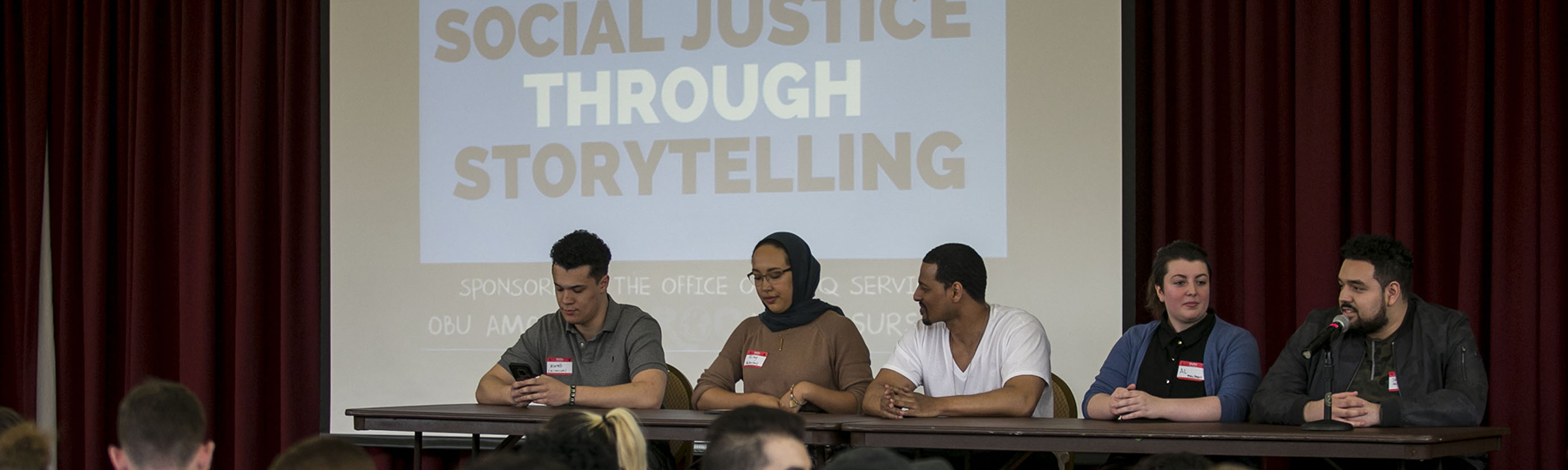 Five panelists are seated at a table in front of a screen that states Social Justice Through Storytelling.
