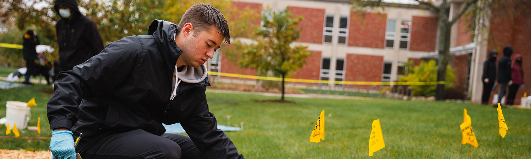 A student wearing a black coat and rubber gloves examines evidence next to yellow flags marking a mock crime scene outside of Anspach Hall.