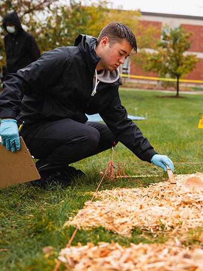 Student wearing black coat and rubber gloves examines evidence next to yellow flags marking a mock crime scene outside of Anspach Hall.