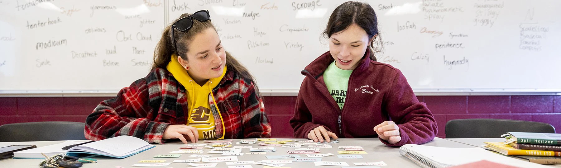 Two creative writing students seated at a table review words on scraps of paper.