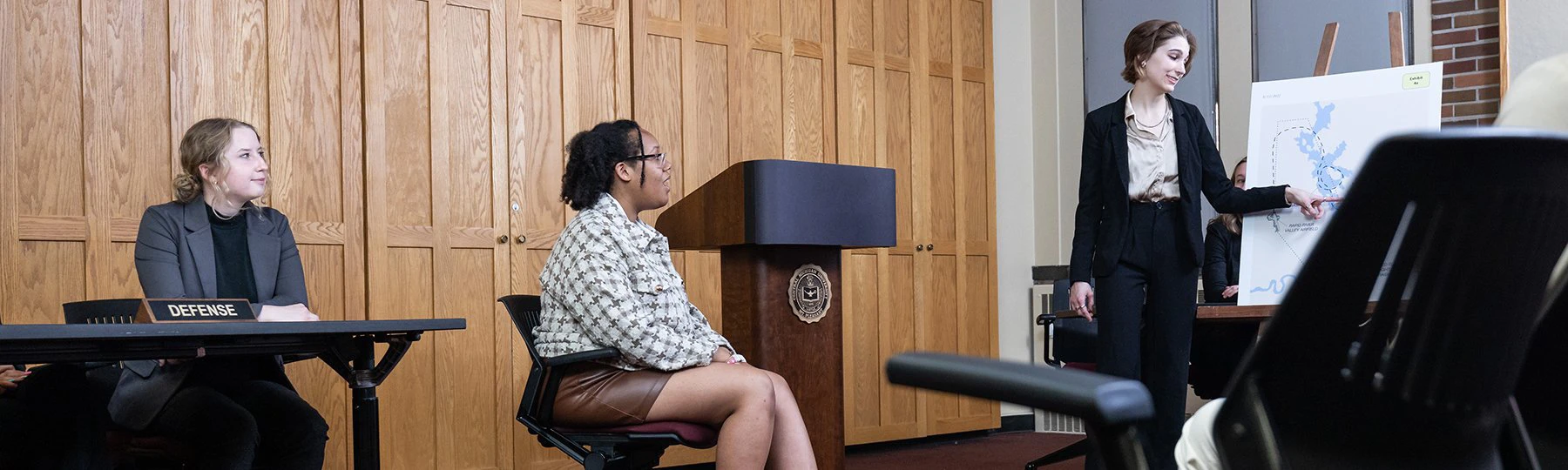 Students seated and standing near a podium question a witness during a mock trial presentation.