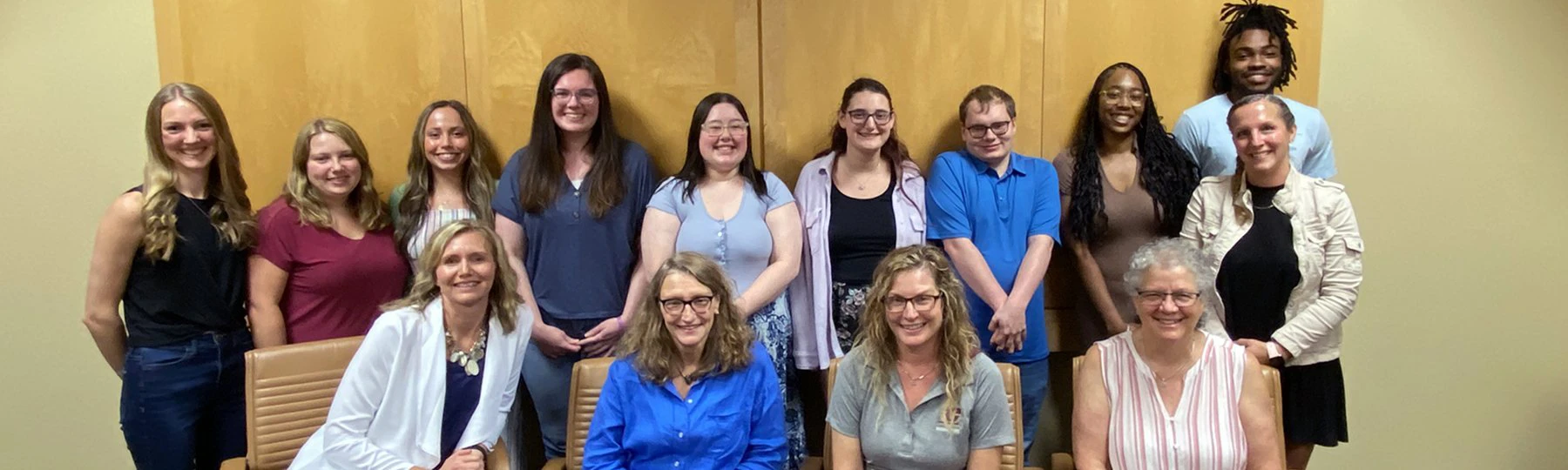 Social Work students stand in front of a wood panel wall behind faculty seated in chairs.