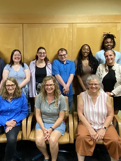 Social Work students stand in front of a wood panel wall behind faculty seated in chairs.