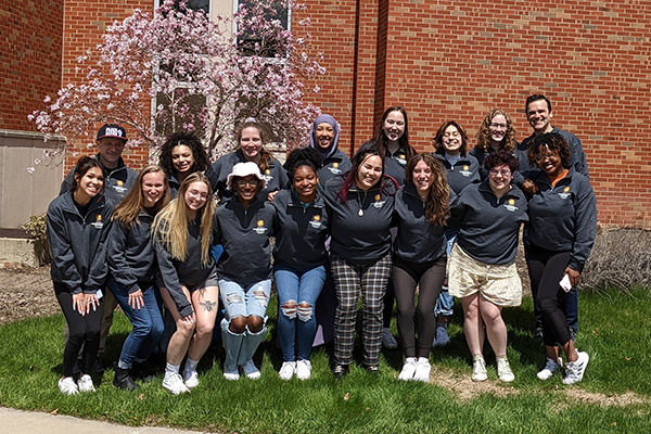 Students and faculty from the Institute for Transformative Dialogue stand together in front of a blossoming tree and a brick building.