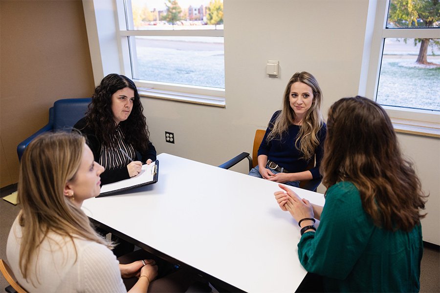 Four women sit around a white table talking. One has a clipboard and pen and is taking notes.
