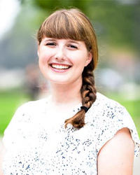A woman smiling at the camera for a professional headshot while outside in front of green trees.