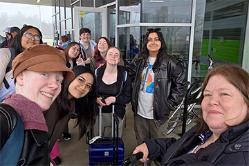 A group of individuals stand next to glass doors with backpacks and rolling luggage bags.