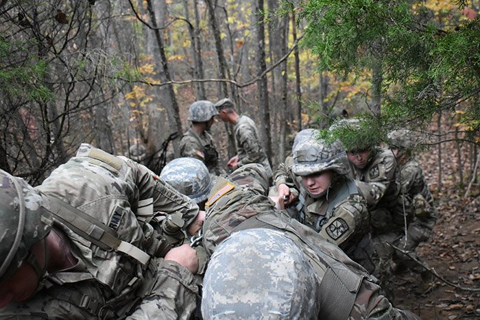 Cadets wearing Army uniforms crawl and crouch on the ground in between trees