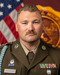 SSG Dustin Houghton wearing his military uniform while seated in front of the American and Central Michigan University flags.