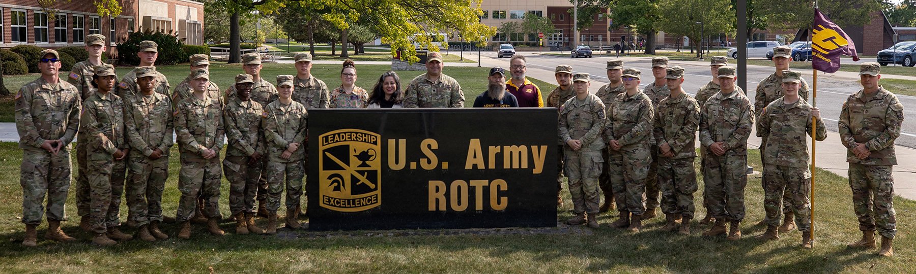 A group of cadets in uniform posing for a photo in front of a Central Michigan University flag.