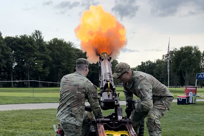Two cadets wearing military uniforms operate a cannon with orange flames and smoke firing out the end