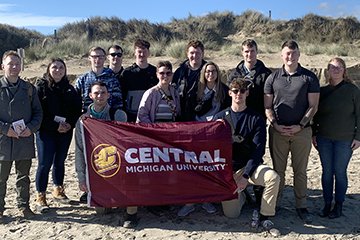Central Michigan University students hold a CMU flag while posing together at Utah Beach in Normandy, France.