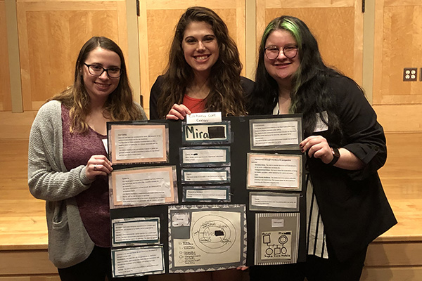 Three Social Work students stand in front of a stage while holding a poster