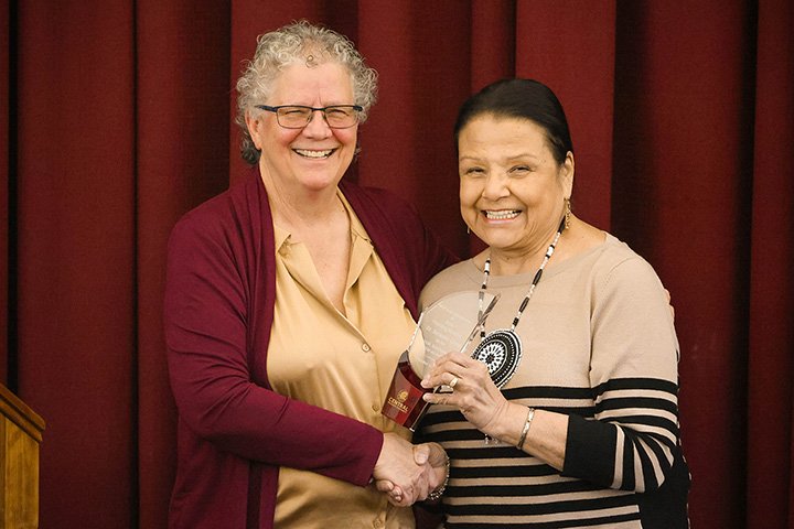 Susan Grettenberger shakes hands with Suzanne Cross while standing in front of a maroon curtain.