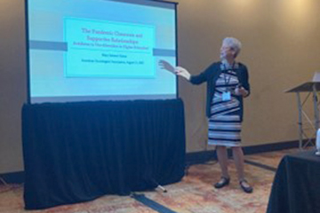 Sociology faculty member Mary Senter wearing striped dress and black sweater stands next to a screen displaying the title of her keynote presentation.