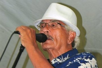Image of Larry T. Reynolds wearing a white hat and blue and white shirt while speaking into a microphone.