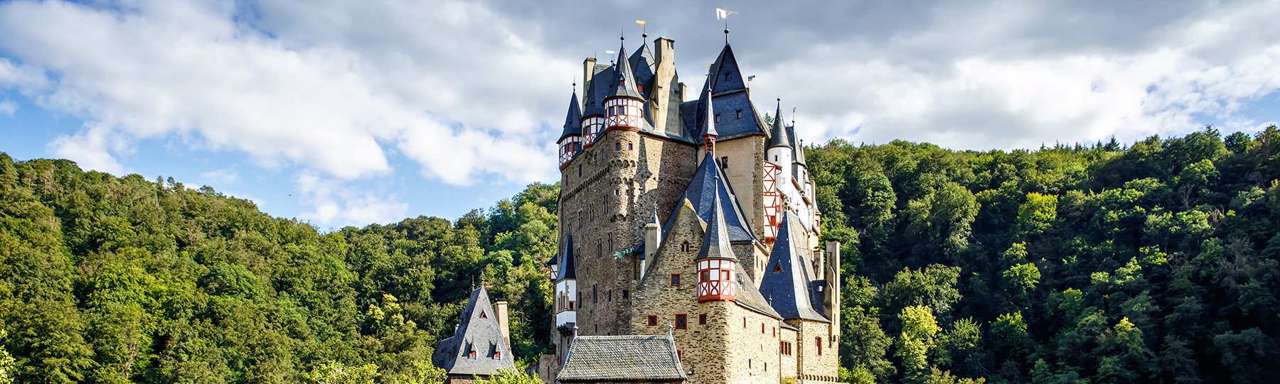 A castle in Germany with multiple towers at the top that are white and red with flags. The castle is sitting in front of a green expansive forest in the background