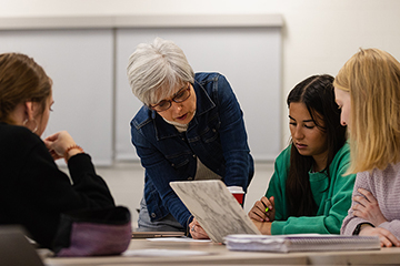 World Languages faculty member Alejandra Rengifo wearing a blue jacket and glasses reviews materials with students seated at tables.