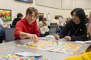 Three individuals seated at a table with a board game and dice in front of them.