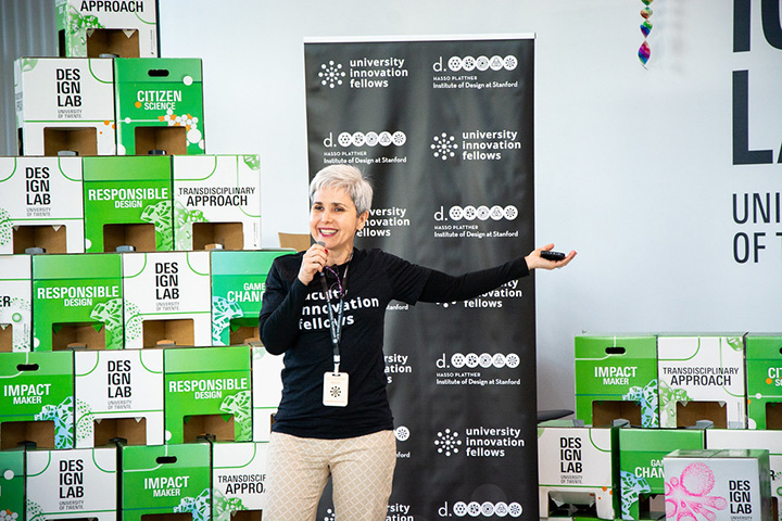 Alejandra Rengifo stands in front of a banner and Design Lab boxes wearing a black shirt and holding a microphone.