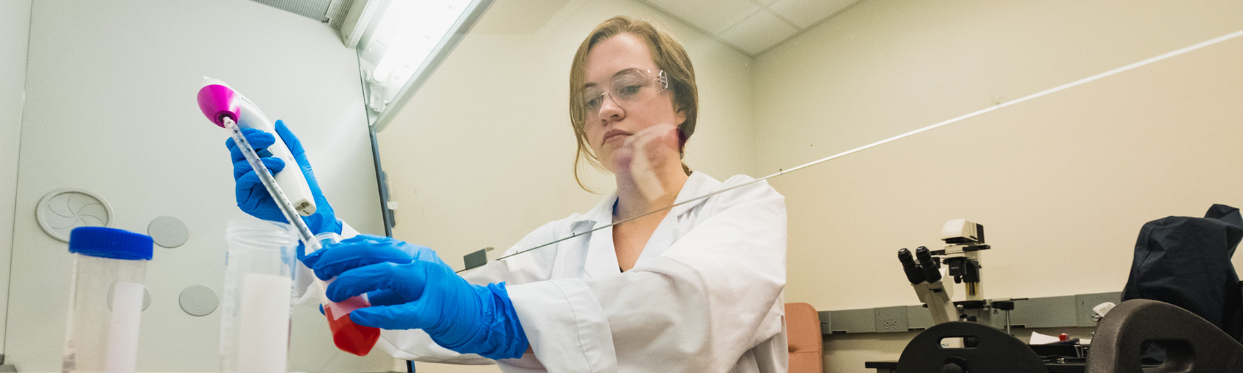 A female student in a lab coat prepares samples under a vent hood in the Sepsis Lab at Central Michigan University.