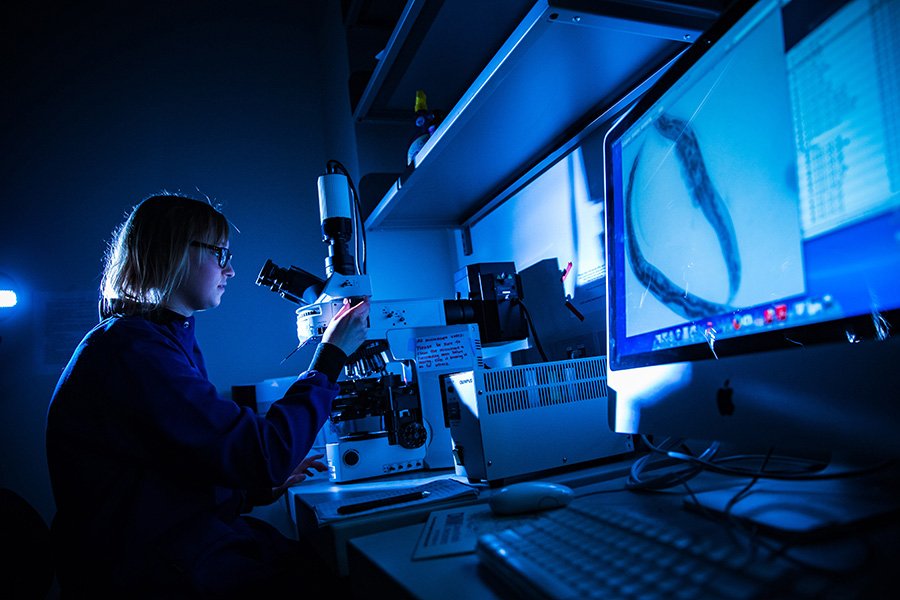 A student in a blue lab coat, wearing glasses, examining a sample in a microscope.