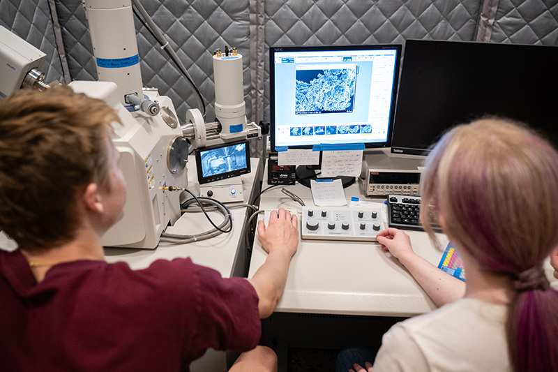2 students sitting in front of a computer and monitor displaying the results from an electron microscope.