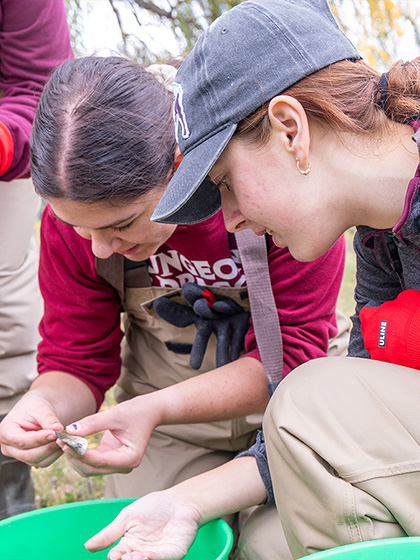 Two students in waders are kneeling examining a small fish