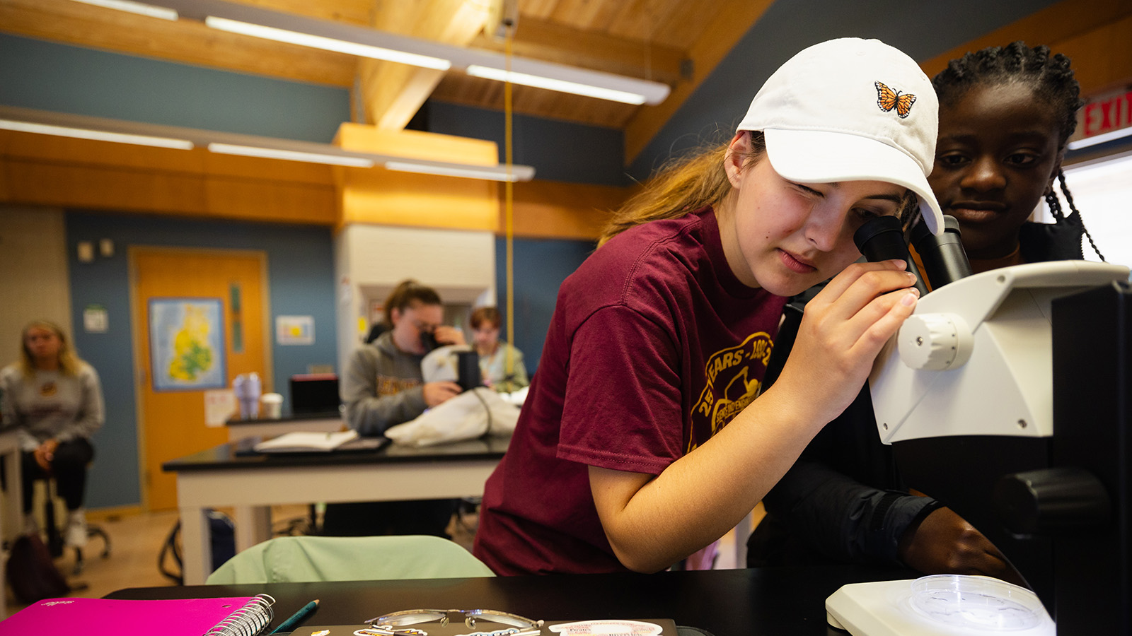 A student in a maroon t-shirt and white hat looking into a microscope.