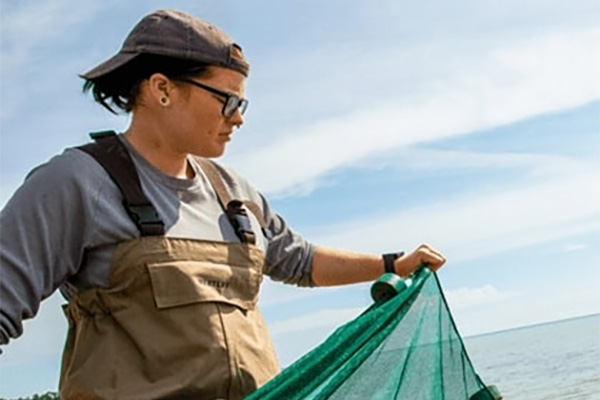 A woman wearing a baseball cap, a gray long sleeve shirt, and brown wader overalls standing in water holding a net
