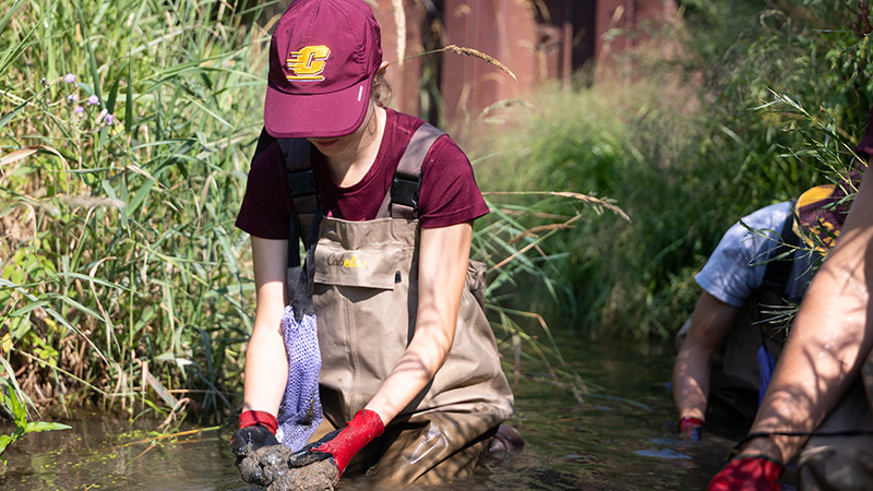 A woman in a maroon hat and waders examining a sample in a muddy body of water.
