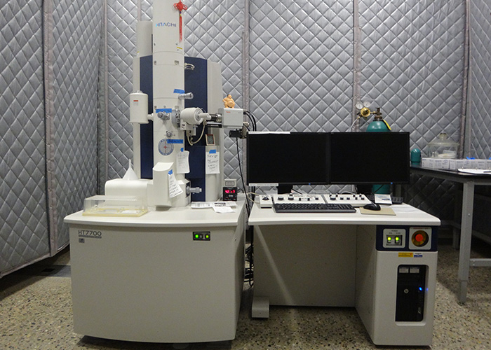 Hitachi HT7700 Scanning Transmission Electron Microscope next to a desk with a computer on it in the microscopy facility.