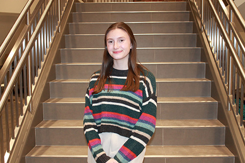 A woman with brown hair wearing a striped shirt standing in front of a staircase.
