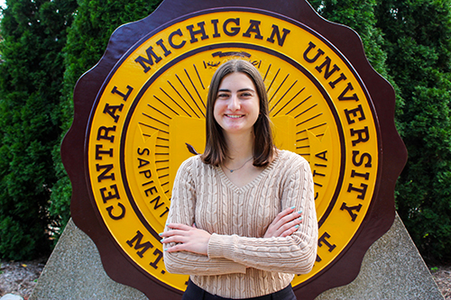 Claire DeBlanc in a tan sweater standing in front of the CMU Seal.