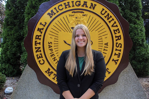 Meghan VanDamme in a green shirt and black jacket in front of the CMU Seal.