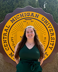 20240516_CS-ZoeyArchbold 200x250 Zoey Archbold in a pale green dress, standing in front of the CMU seal, smiling at the camera.