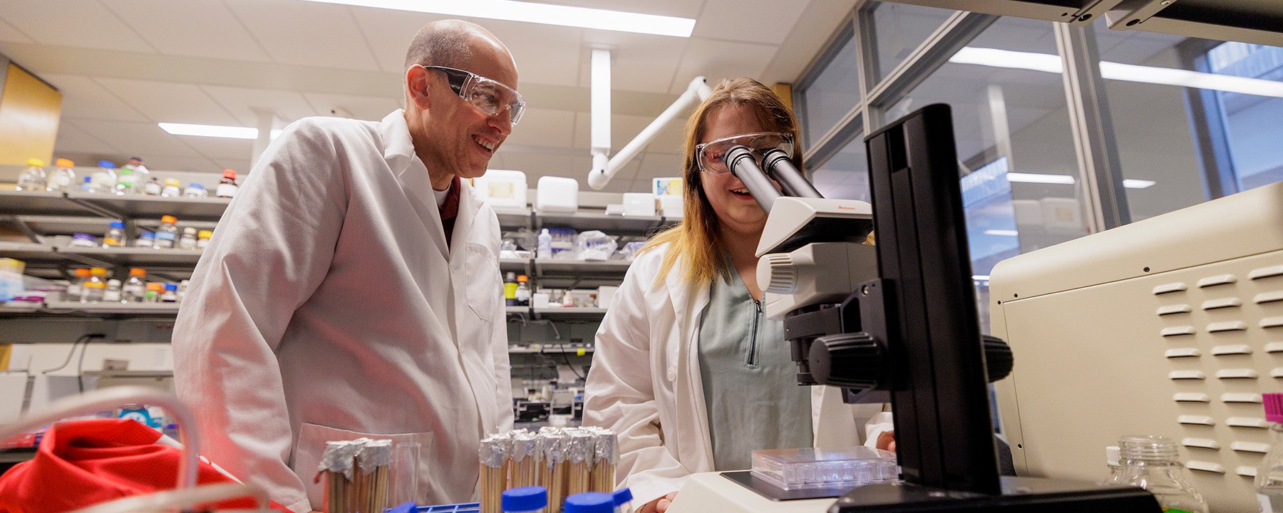 A faculty member and student in safety glasses and lab coats examining something under a microscope.