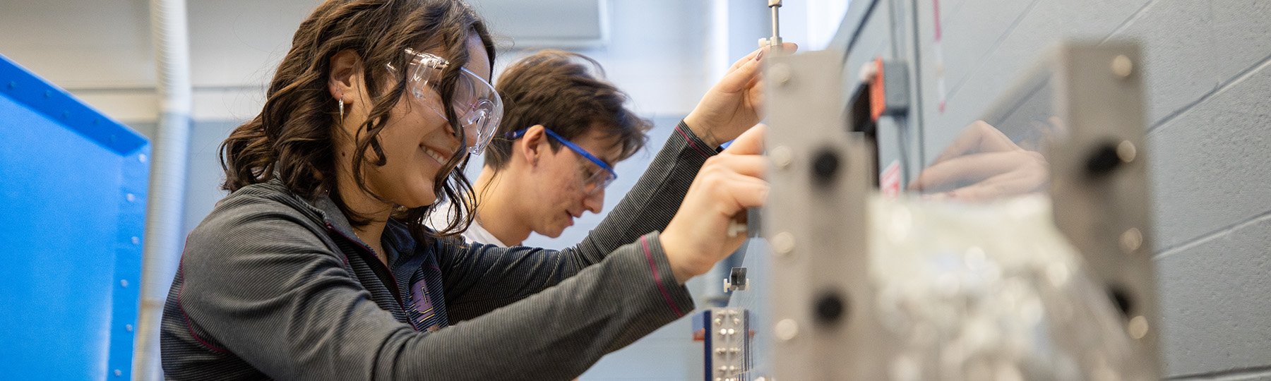 Two students wearing safety goggles work together on a hands-on engineering project in a lab setting.