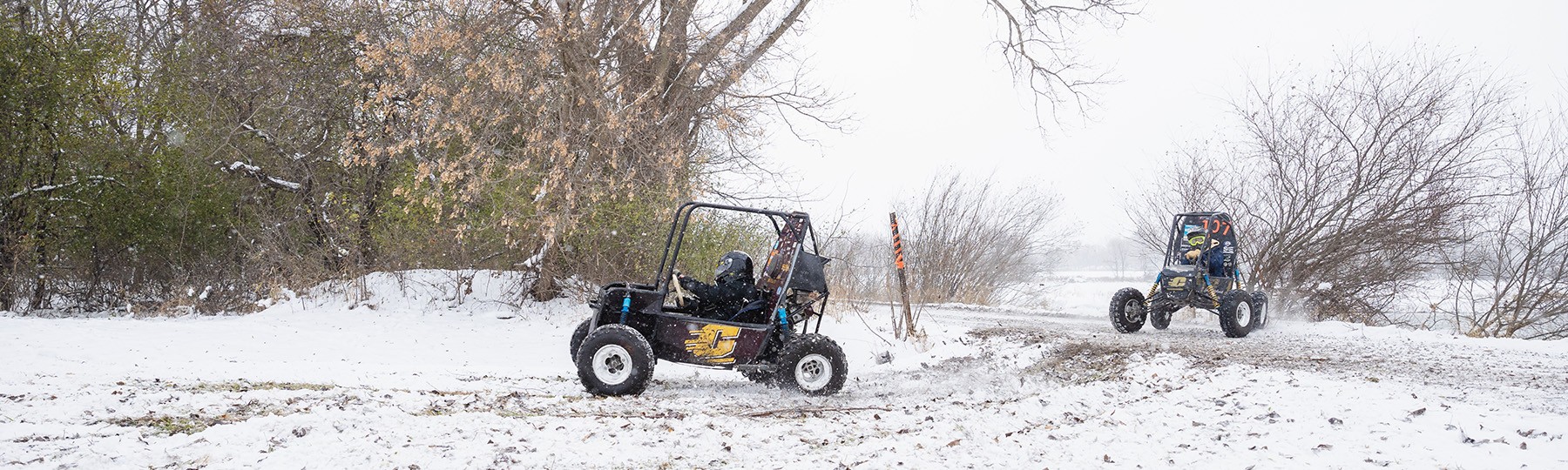 Two student-built baja cars driving on an off-road course on a snowy day.