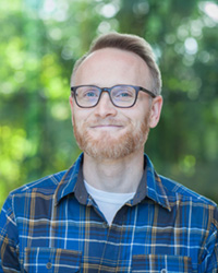 A man stands outside in blue flannel shirt and glasses.