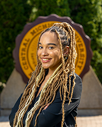 Mya Grant in a dark outfit standing in front of the Central Michigan University seal.