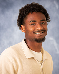 A man with a goatee and beard in a beige shirt in front of a grey backdrop.