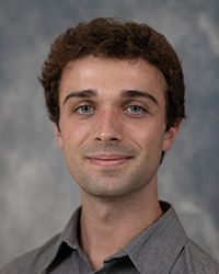 A close-up of a man with dark hair in a grey shirt against a grey background.